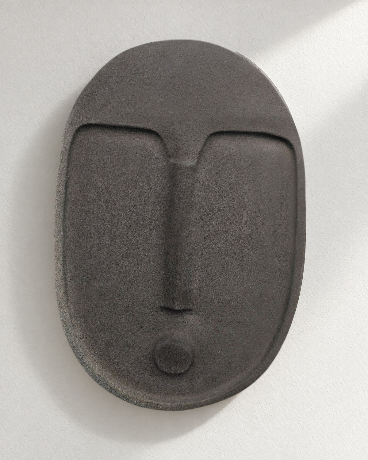 The anthracite ceramic wall mask, front view, on a pale plaster wall in soft window light