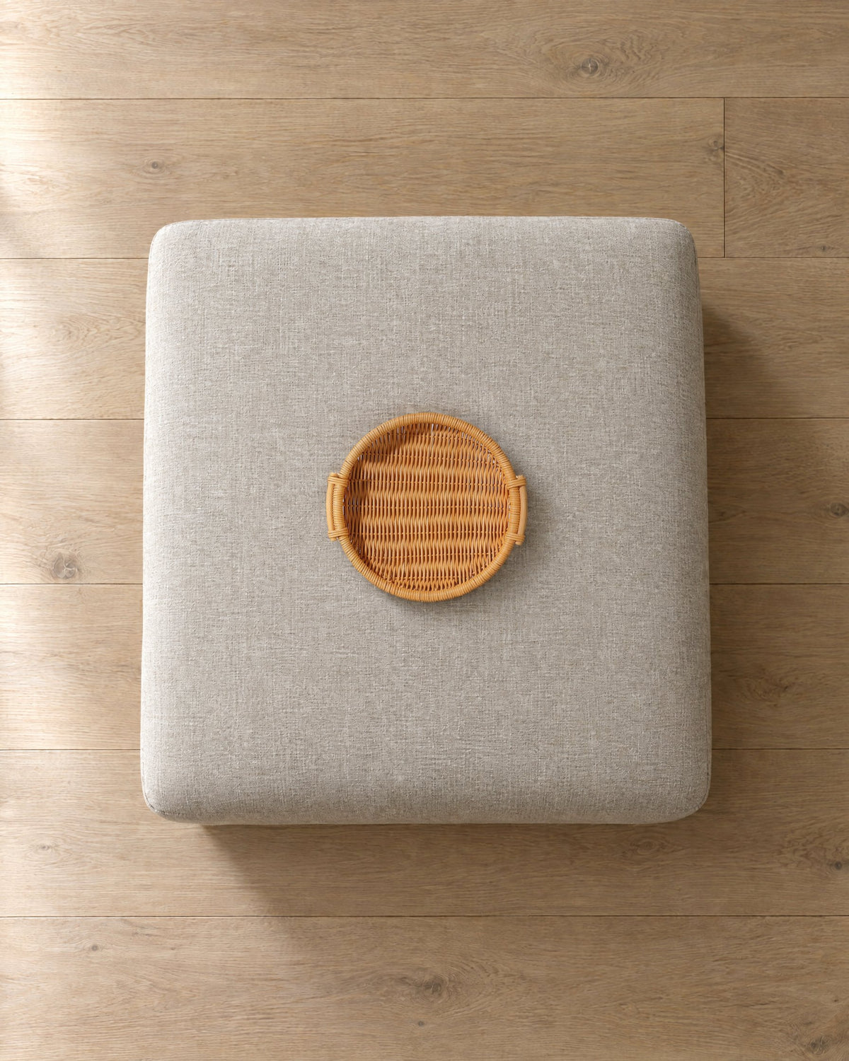 Round honey woven rattan tray, 10 in across, on a 30 in square oatmeal linen ottoman, seen from above