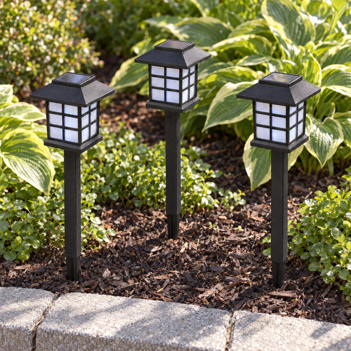 Three black pagoda solar stake lights, switched off, standing in a mulched flower bed among hostas in daylight